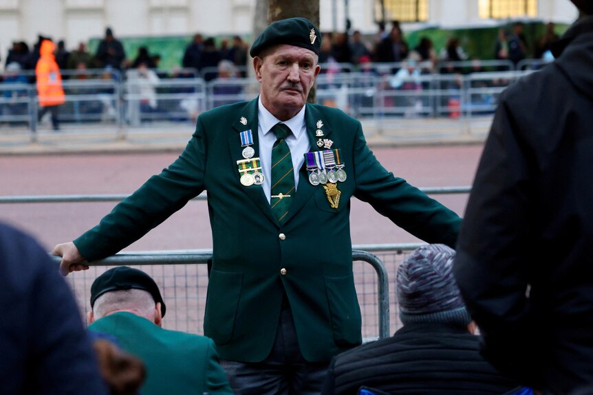A man in green blazer and beret with war medals leans against a barricade. 