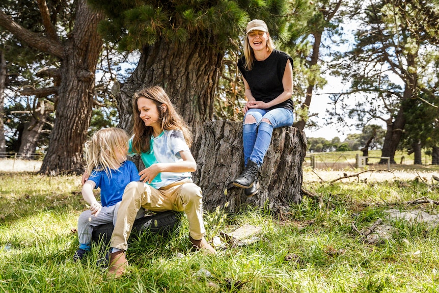 Two boys wrestle on an old tyre while a woman sitting on a log in the background laughs watching.