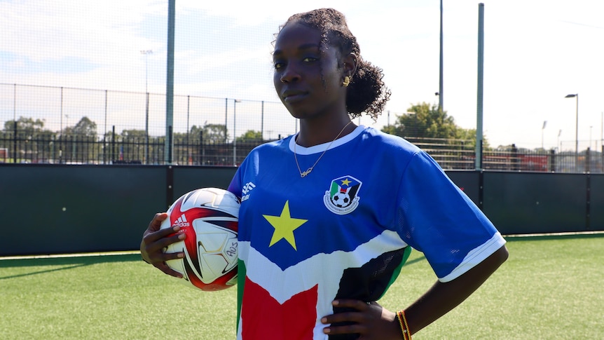 A girl stands with a soccer ball, in a South Sudan uniform.