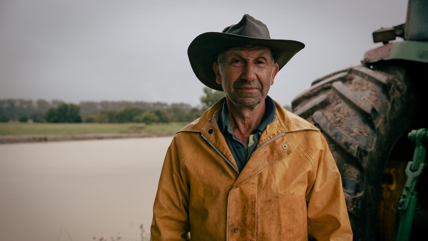 An old man wearing a yellow PVC jacket and brown akubra stands in front of a tractor tyre and brown water dam.