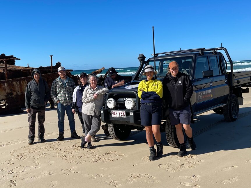 seven people stand in front of a ute on sand with the ocean and a rusty shipwreck in the background