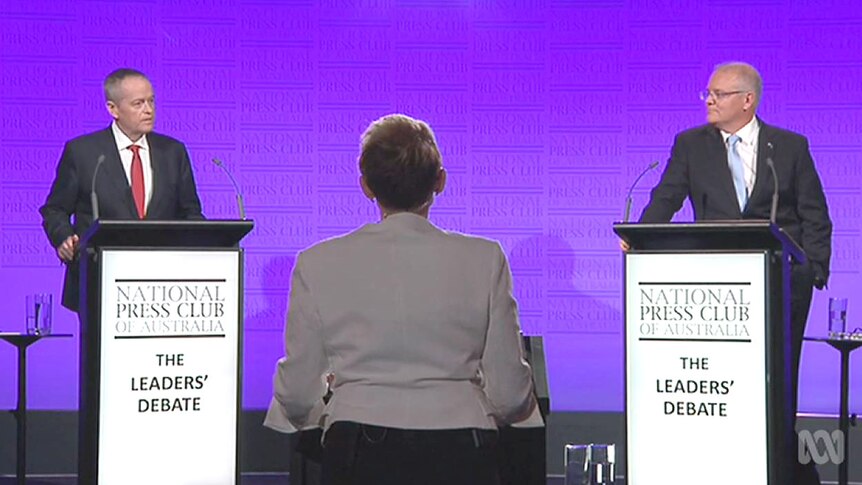 The two men at podiums, with purple background and moderator in the middle.