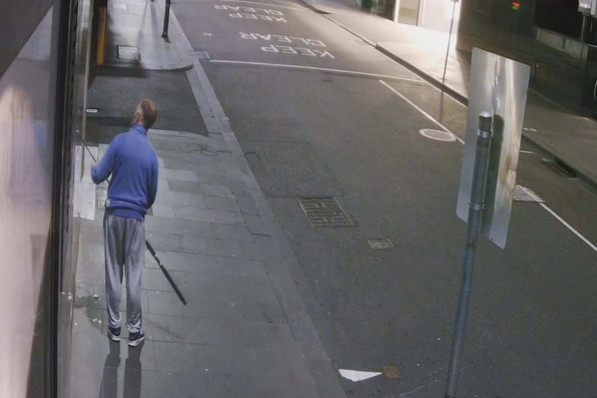 A still from CCTV vision of a man pushing a fishing rod through a shopfront window, wearing a blue jacket.