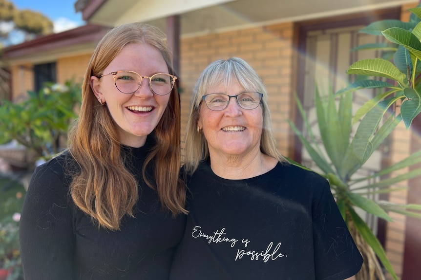 A teenage girl and a woman stand close together, both smiling, in the backyard of a home