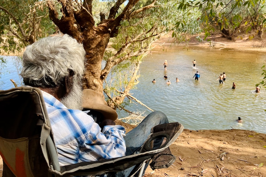 An old man with white hair sits by a river bank with a dozen children visible in river behind.