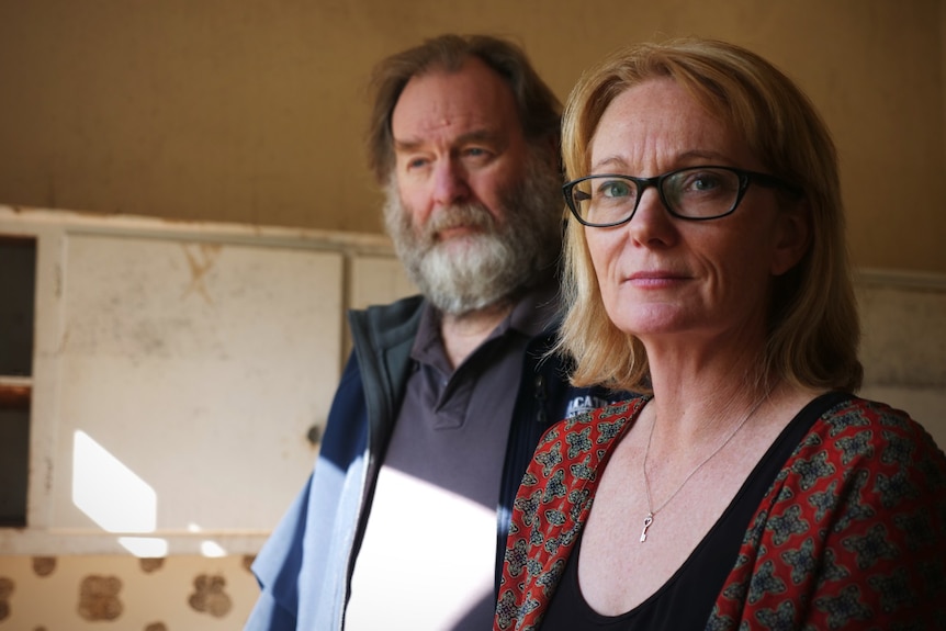 A woman with blonde hair and glasses and a man with a bear in a flood-damaged house