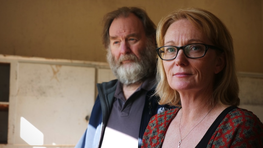 A woman with blonde hair and glasses and a man with a bear in a flood-damaged house