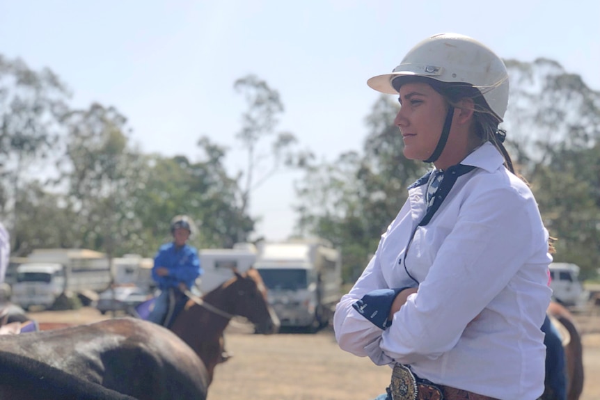 A teenage girl sitting on top of a horse, not looking at the camera. Her arms are crossed and she's pondering in the distance.