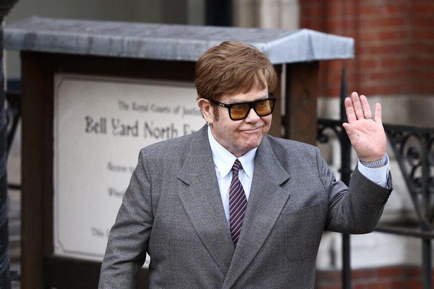 Elton John wears grey suit, white shirt and tie, waves with left hand as he looks to his left. 