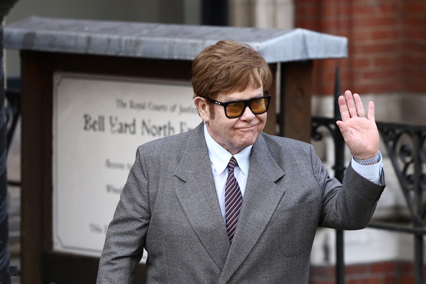 Elton John wears grey suit, white shirt and tie, waves with left hand as he looks to his left. 