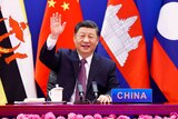 Sitting in front of several flags, Xi Jinping waves to the camera, with a placard stating "China" on th edesk in front of him