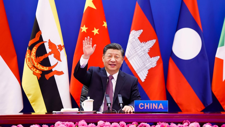 Sitting in front of several flags, Xi Jinping waves to the camera, with a placard stating "China" on th edesk in front of him