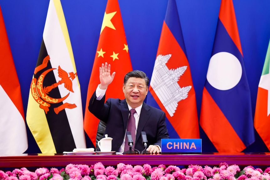 Sitting in front of several flags, Xi Jinping waves to the camera, with a placard stating "China" on th edesk in front of him