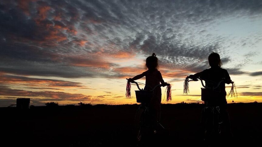 Two silhouetted children ride their bikes into the sunset, the ribbons on the end of their handle bars visible.