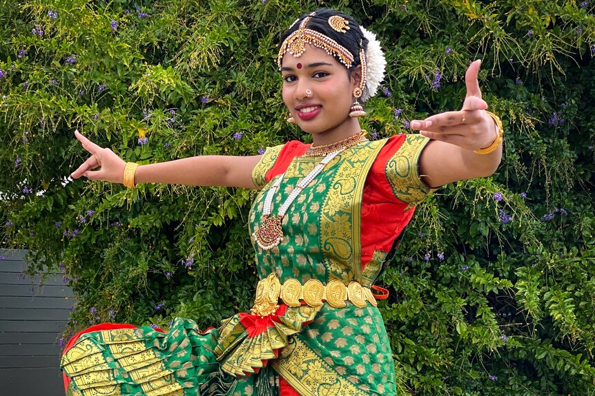 A young brown woman dressed in green and orange sari makes hand gestures, as part of an Indian classical dance.