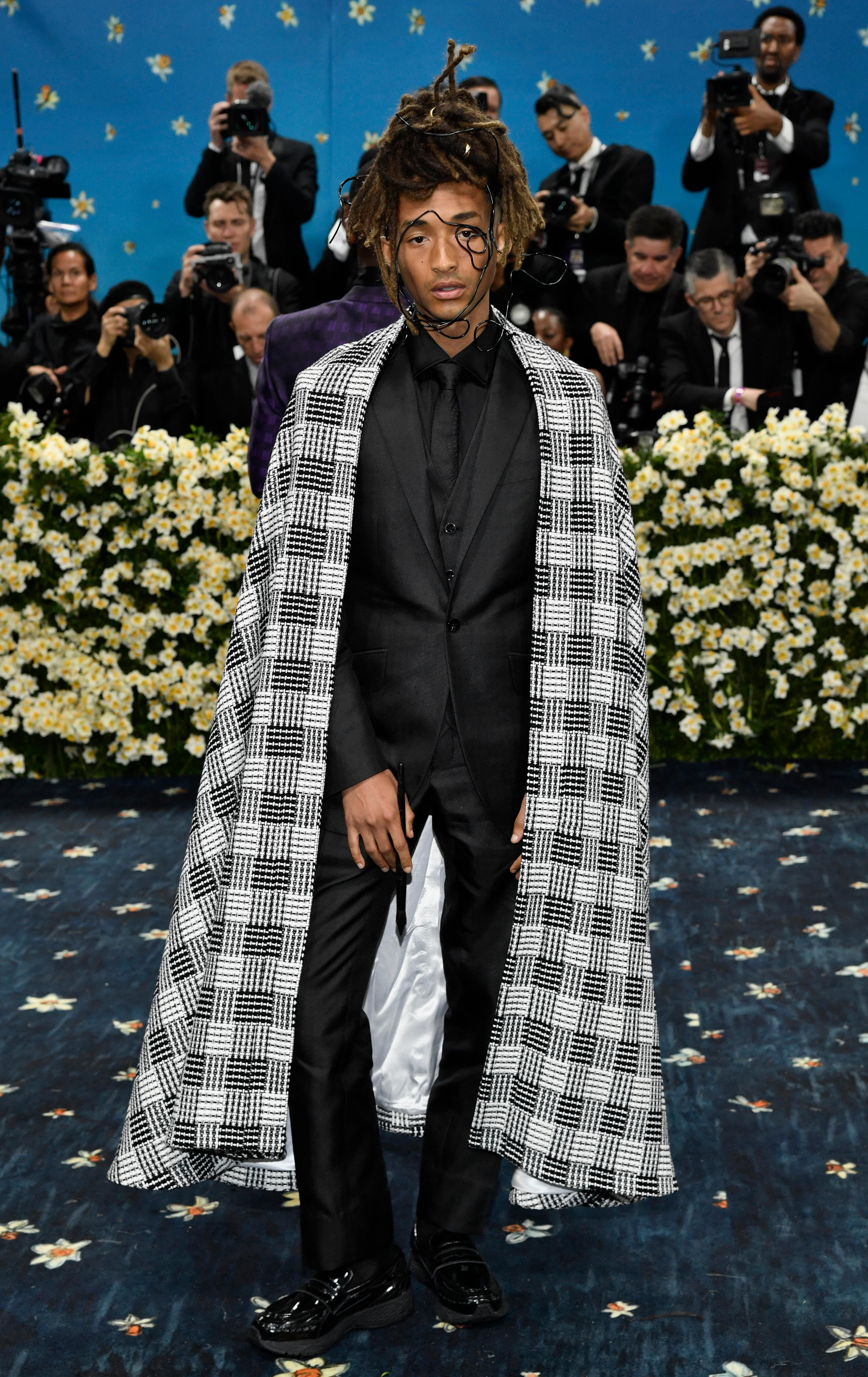 Jaden Smith wearing a black suit with a black and silver checked cape and black wire coiled over his face 