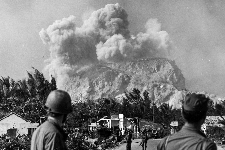 Soldiers looking at large smoke and dust plume on top of mountain in the distance.
