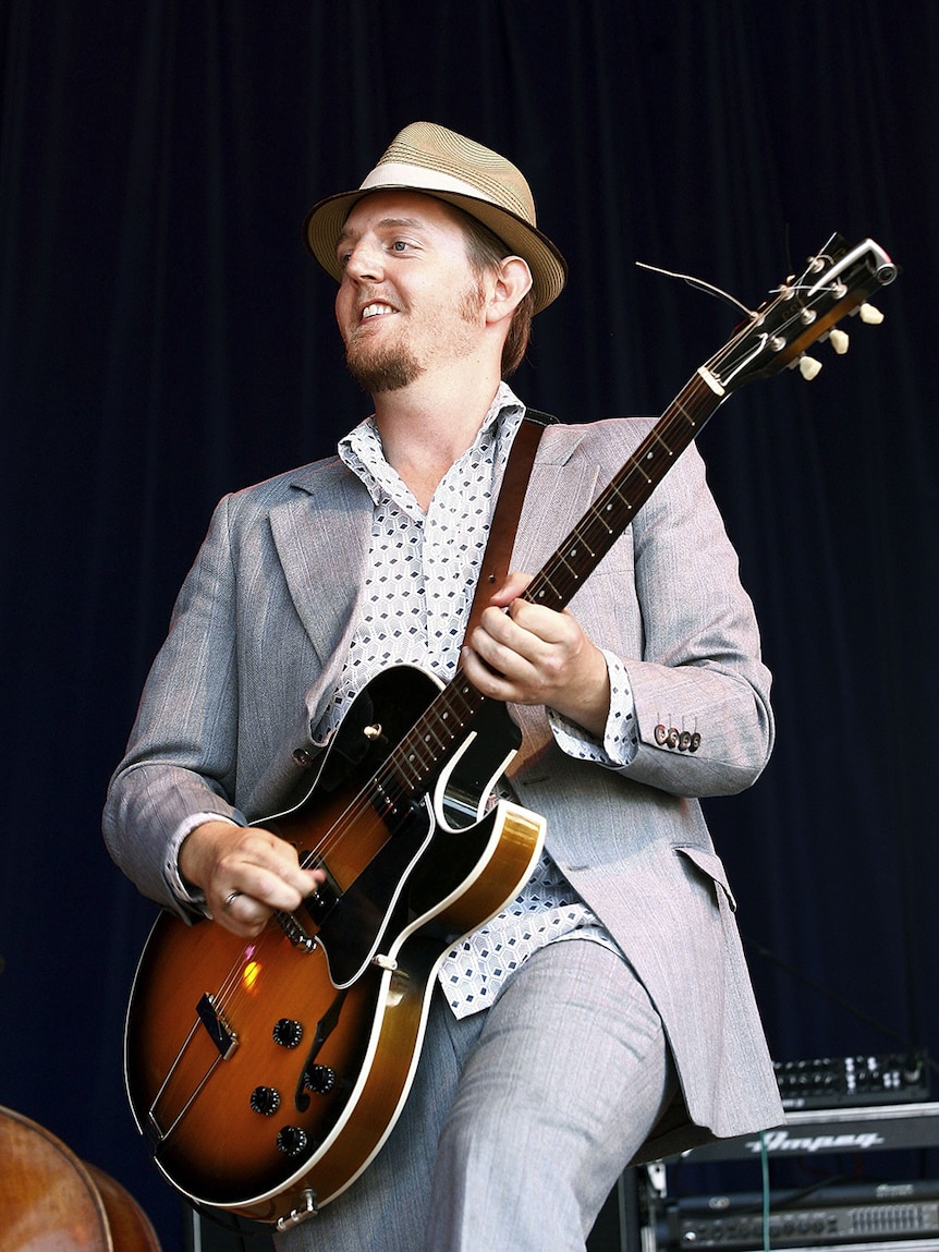 Tristan Goodall of The Audreys plays guitar on stage. He smiles and wears a hat and grey suit jacket.
