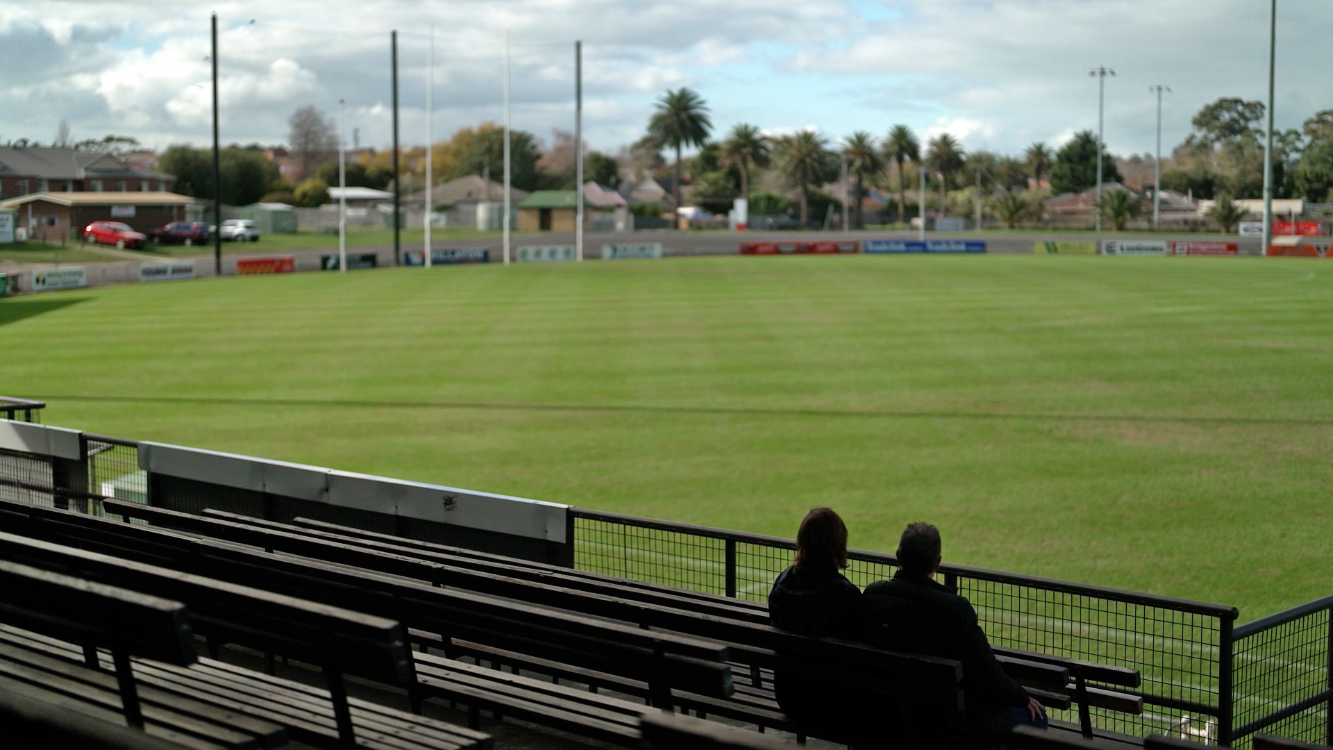 Two people silhouetted sit in the stands of sports field.