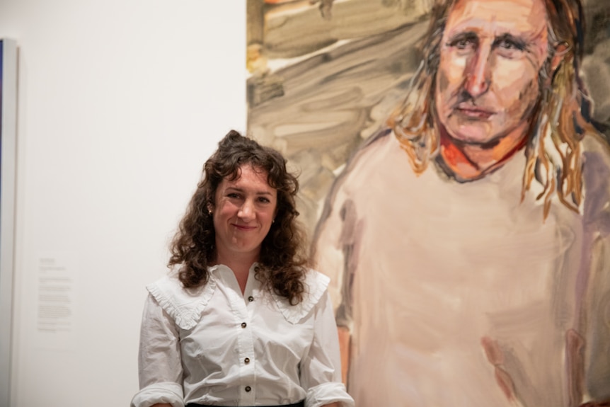 A woman in her early 40s - the artist Laura Jones - smiling and standing in front of her painting of Tim Winton