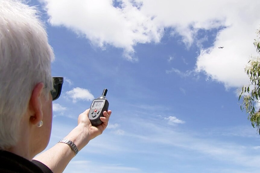 A woman watches a plane fly overhead while holding out a noise-measuring device.