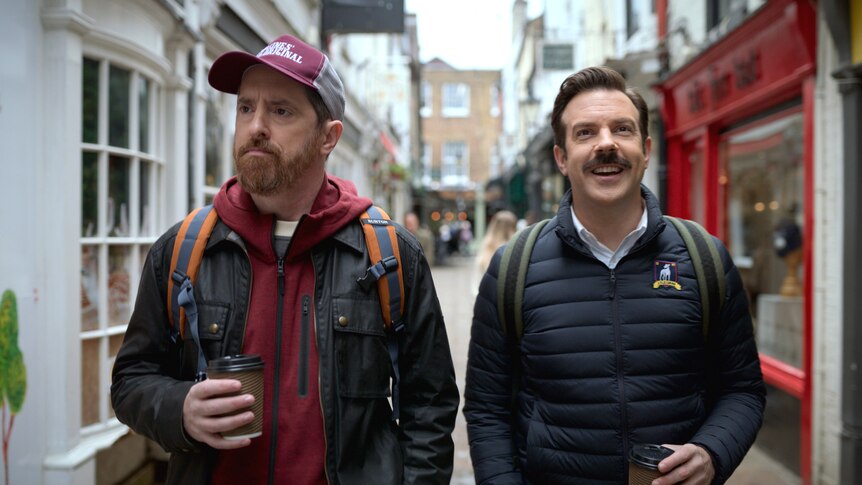 Jason Sudeikis (Ted) and Brendan Hunt (Beard) talk while holding takeaway coffees in a scene from Ted Lasso.
