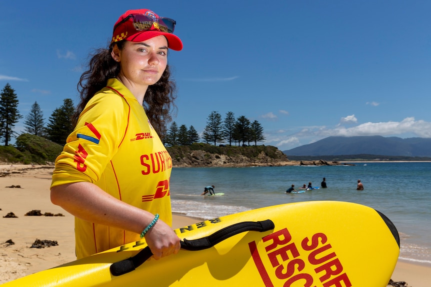 A young woman in surf life saving uniform on the a sunny beach while holding a club board 