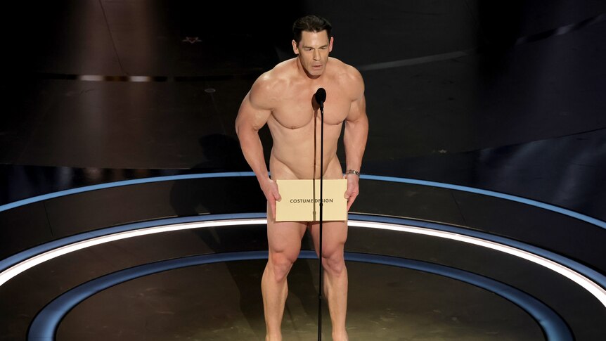 A man stands on stage naked holding an envelope over his groin. He's talking into a microphone