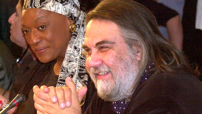 A man with brown hair and a grey beard sits alongside a woman wearing a scarf  