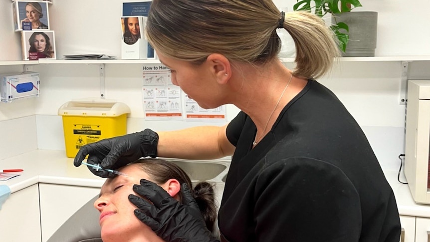 Woman wearing black t-shirt and black gloves, inserts a needle near the eye of a woman lying down