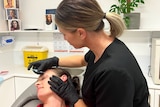 Woman wearing black t-shirt and black gloves, inserts a needle near the eye of a woman lying down