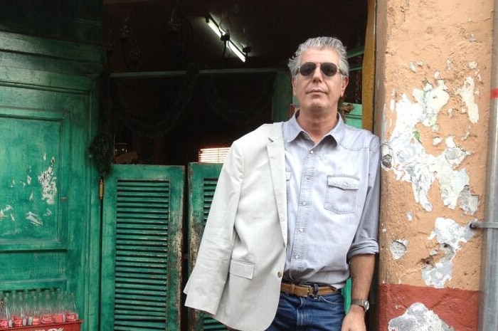 Anthony Bourdain stands in a doorway with peeling orange paing, wearing a jacket slung over one shoulder and sunglasses