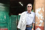Anthony Bourdain stands in a doorway with peeling orange paing, wearing a jacket slung over one shoulder and sunglasses