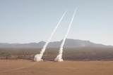 Two missiles launch with a cloud trail from the desert.