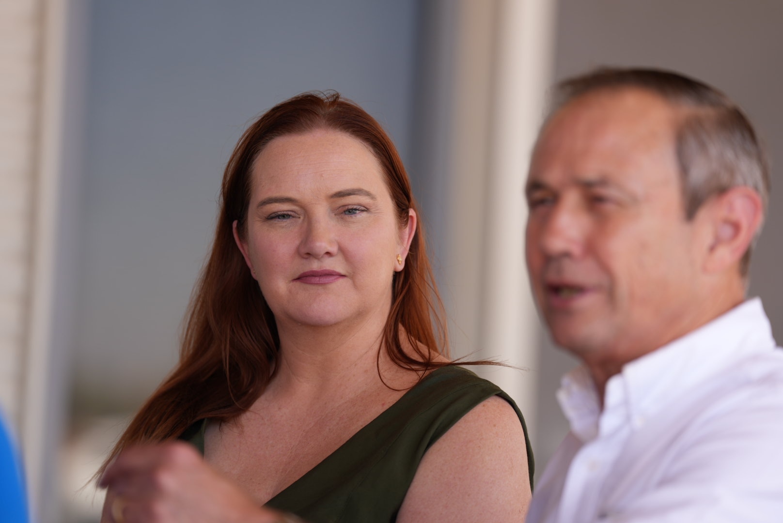 A woman with red hair looks on as an older man in white shirt addresses reporters.
