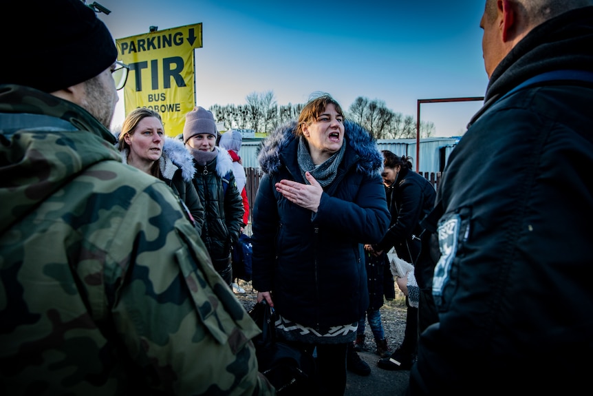 A woman in a fur trimmed coat gesticulates to men in camo gear 