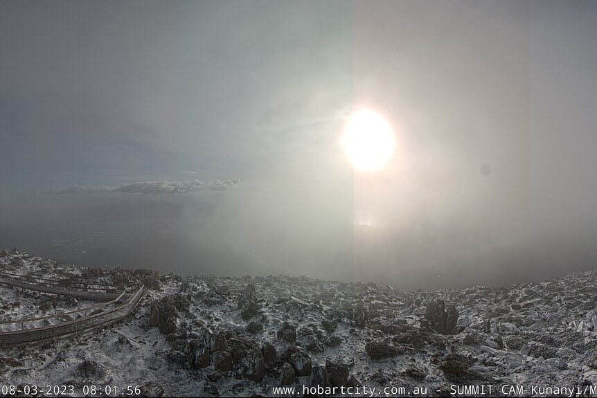 A webcam image showing snow covered rocks and the sun obscured by clouds.