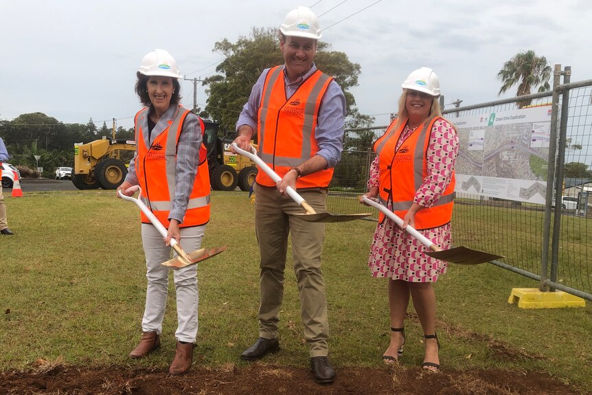 Man standing between two women, all wearing orange high-vis vests and holding shovels with a hole in the dirt in front of them