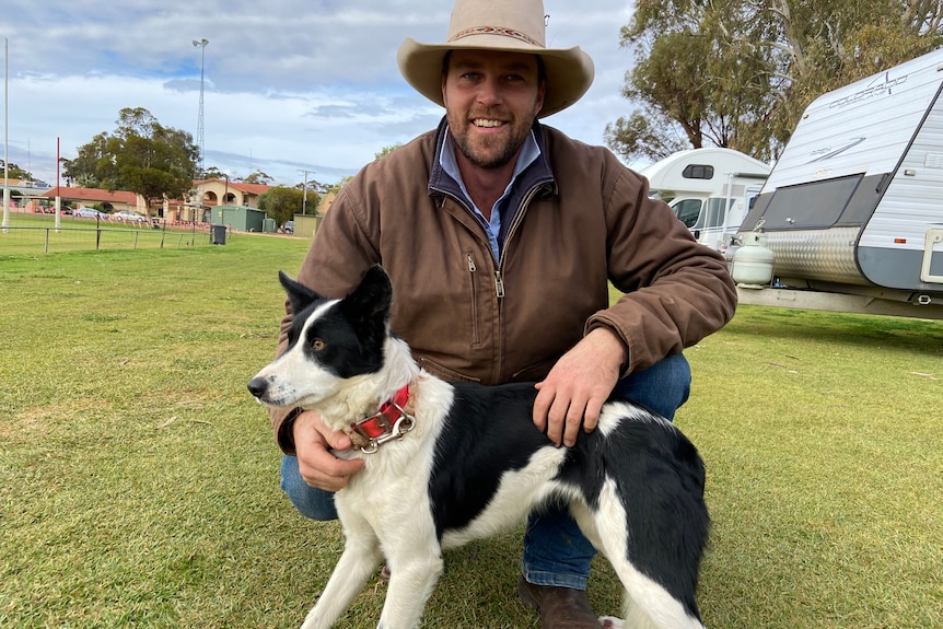 A man wearing a hat kneels next to a border collie on an oval, caravans in the background.