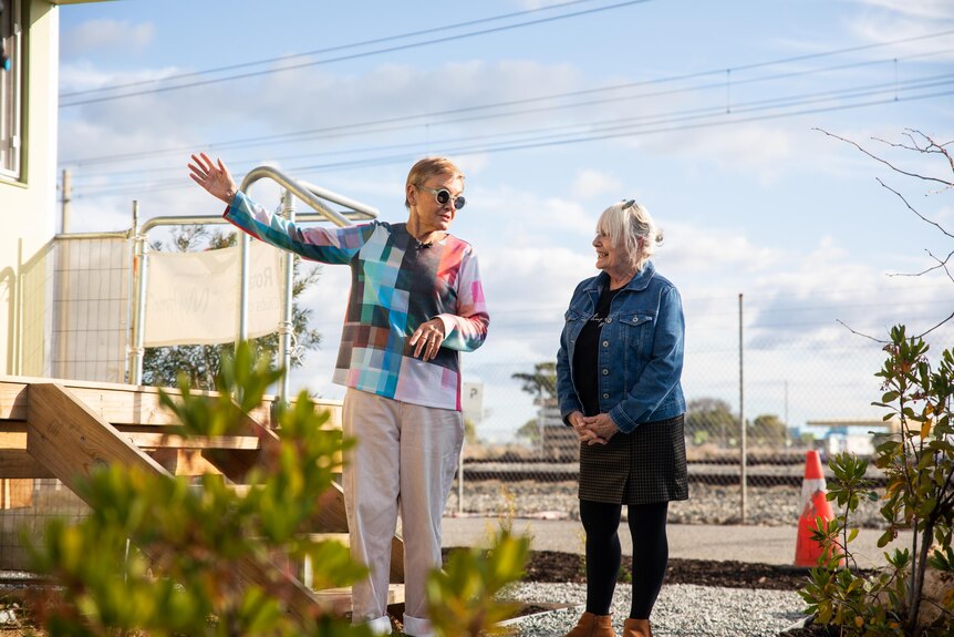 A woman in a colourful shirt gestures at a build while talking to a woman in a denim jacket next to her.