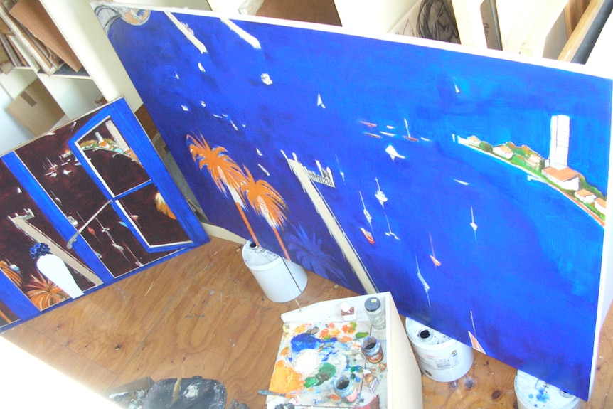 A shot from above of a large blue painting of a harbour in progress with a smaller work of a harbour through a window next to it