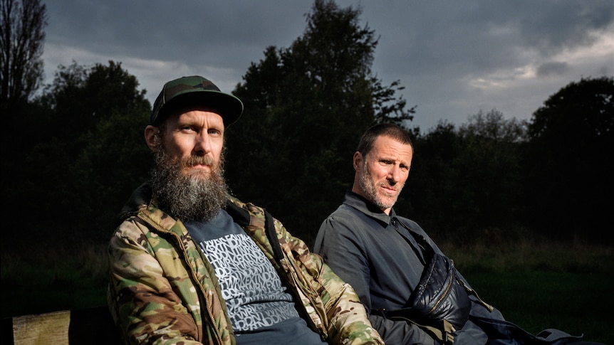 Two men, the members of Sleaford Mods, sit outdoors and glare at the camera