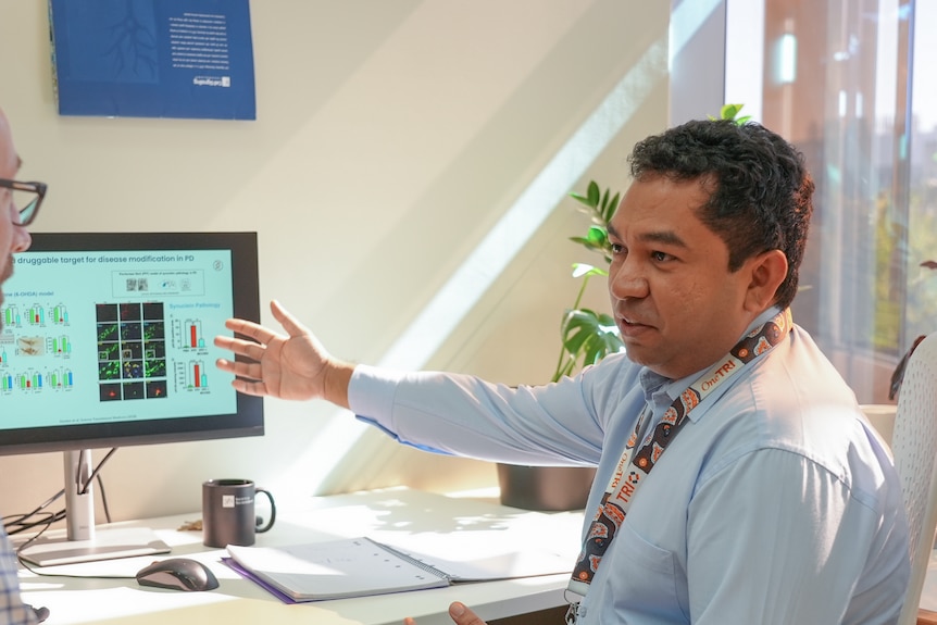 A man sitting at a desk gestures at graphs on a computer screen.