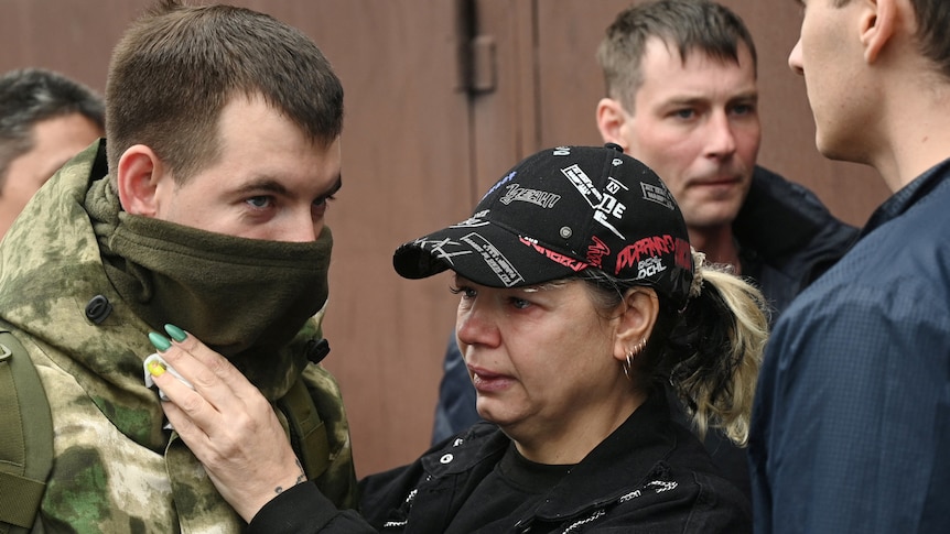 A woman in a baseball cap looks teary while adjusting a young man's military uniform