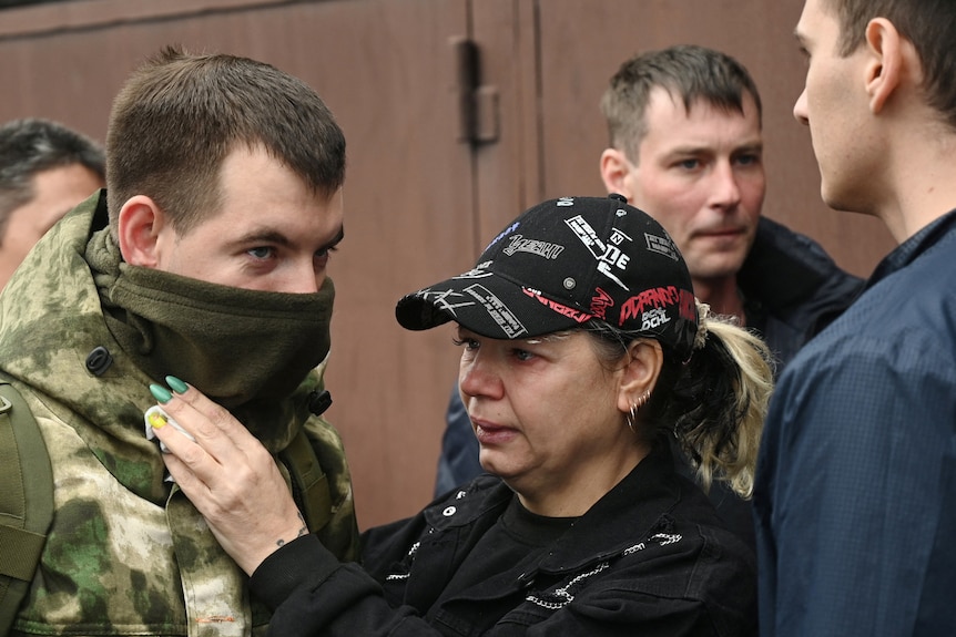 A woman in a baseball cap looks teary while adjusting a young man's military uniform