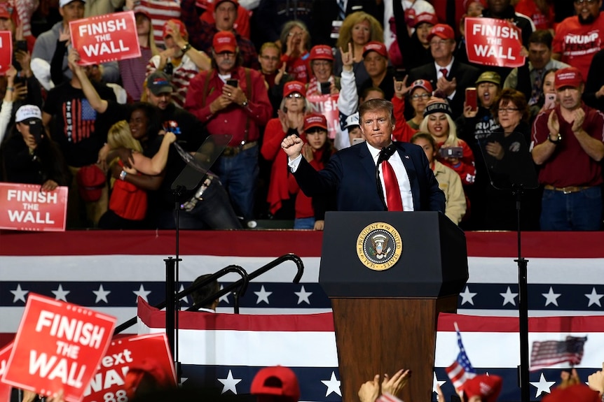 A man in a suit holds his fist up as he speaks to a large crowd waving signs in support.