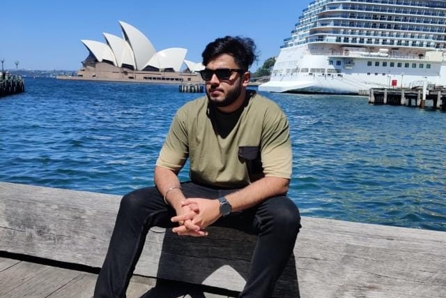 Man in sunglasses sits in front of Sydney harbour overlooking Sydney Opera House