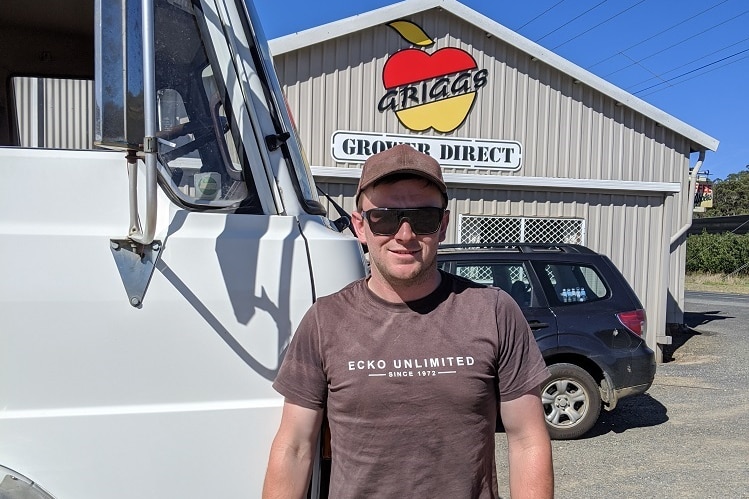 A man smiles as he stands next to the cab of a truck parked in front of a shed signposted 'Griggs grower direct'.