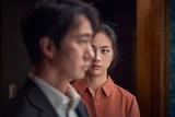  An East Asian woman in a brown top stares intently at an East Asian man. He is out of focus, and wearing a black suit.
