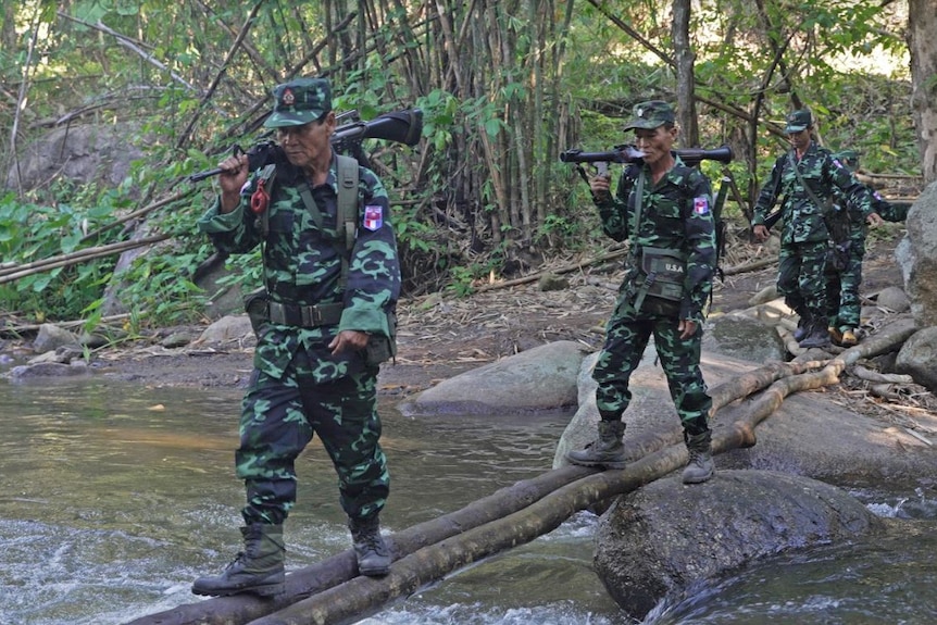 Three people dressed in army fatigues walk along a log over a river while holding guns.